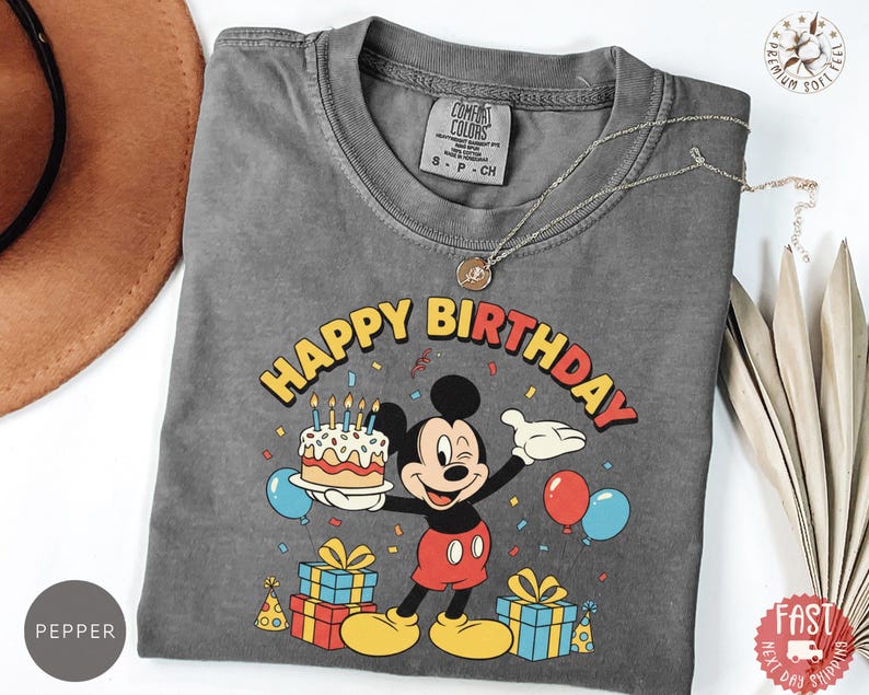 K&ouml;nnte beinhalten: Ein graues Comfort Colors T-Shirt mit einer bunten Grafik von Mickey Mouse, die eine Geburtstagstorte, Luftballons und Geschenke h&auml;lt. Die Worte "Happy Birthday" sind in Gelb und Rot gehalten. Eine silberne Halskette liegt &uuml;ber dem Shirt.