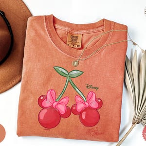 Peut inclure: T-shirt corail avec un motif de cerises Minnie Mouse. Le graphisme comprend deux cerises avec des n&oelig;uds roses et des feuilles vertes, avec les logos Disney et Minnie Mouse. Un collier en or est drap&eacute; sur le t-shirt.