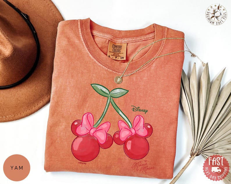Peut inclure: T-shirt corail avec un motif de cerises Minnie Mouse. Le graphisme comprend deux cerises avec des n&oelig;uds roses et des feuilles vertes, avec les logos Disney et Minnie Mouse. Un collier en or est drap&eacute; sur le t-shirt.