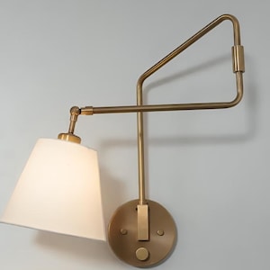 Könnte beinhalten: Eine Wandleuchte aus Messing mit einem weißen Stoffschirm. Die Leuchte hat ein geometrisches Design mit einem langen Arm, der sich von der Wand erstreckt, und einem kürzeren Arm, der den Schirm hält.