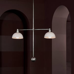 Puede incluir: Una lámpara colgante moderna de dos luces con acabado metálico cepillado. La lámpara presenta dos pantallas blancas en forma de cúpula y una barra vertical central con una esfera decorativa. El diseño es minimalista y adecuado para varios estilos de interior.