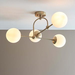 Può includere: Una lampada da soffitto con struttura in ottone spazzolato e quattro globi sferici in vetro bianco satinato. La struttura è composta da una piastra di montaggio centrale con un braccio curvo che sostiene le sfere di vetro illuminate.