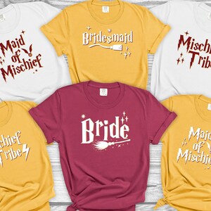 May include: A collection of t-shirts in various colors, including white, yellow, and burgundy. Each shirt features a unique white graphic design with text such as "Bride", "Bridesmaid", and "Mischief Tribe". The shirts are likely for a bridal party.