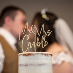 Könnte beinhalten: Ein Kuchenaufsatz aus Holz mit der Aufschrift "Mr & Mrs Grable" in Schreibschrift, platziert auf einem weiß glasierten Kuchen. Der Hintergrund zeigt ein verschwommenes Paar, was auf eine Hochzeitsfeier hindeutet.