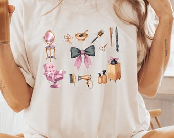 May include: Cream-colored t-shirt featuring a collection of beauty-related illustrations, including a pink salon chair, a large bow, and various hair styling tools. The shirt is worn by a person sitting cross-legged.