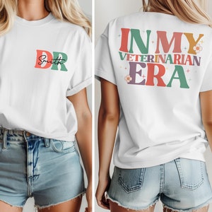 May include: White t-shirt with a colorful "DR Smith" design on the front and "IN MY VETERINARIAN ERA" on the back. The shirt is being modeled by a person wearing denim shorts.