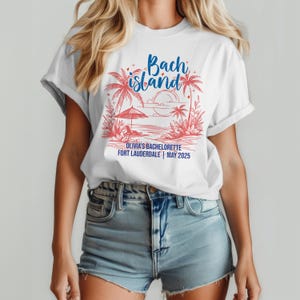 May include: White t-shirt with a beach-themed graphic in red and blue. The design includes palm trees, an umbrella, and the words "Bach Island." Below the graphic, it reads "Olivia's Bachelorette Fort Lauderdale | May 2025."