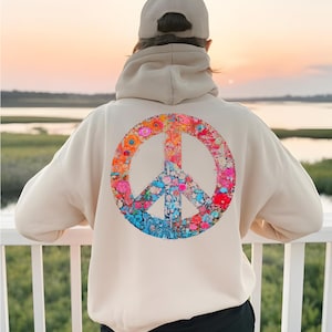 May include: A cream colored hoodie with a large peace sign on the back. The peace sign is made up of colorful flowers and is a vibrant orange, pink, blue, and green.