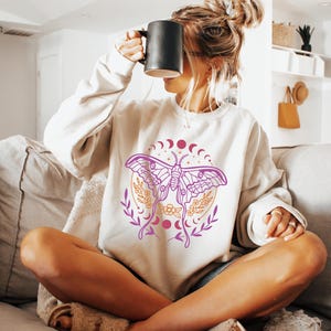 May include: A white sweatshirt with a pink and gold moth design. The moth is surrounded by a circle of moons and stars. The sweatshirt is worn by a person sitting on a couch.