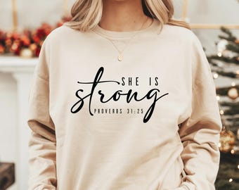 Könnte beinhalten: Ein beiger Pullover mit dem Text "SHE IS strong" und "PROVERBS 31:25" in Schwarz gedruckt.