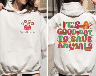 May include: White hoodie with a graphic on the front that says "Dr. Olivia" and a cartoon dog bowl with a bone. The back of the hoodie has a graphic that says "It's a good day to save animals" with a variety of colorful cartoon dog bowls and bones.