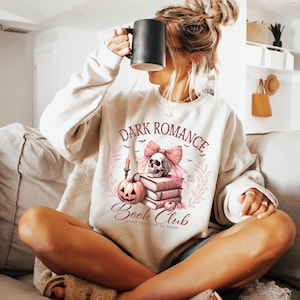 May include: A white sweatshirt with a pink and black graphic design featuring a skull, a pumpkin, and books. The text on the sweatshirt reads "Dark Romance Book Club Always Falling for the Villain".