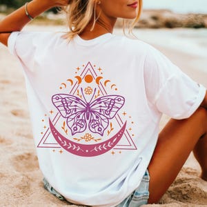 May include: White t-shirt with a purple butterfly graphic design. The butterfly is surrounded by a crescent moon, stars, and a triangle.