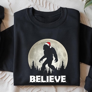 May include: Black sweatshirt featuring a Bigfoot silhouette wearing a Santa hat, set against a full moon and forest. The word "BELIEVE" is printed in white below the image. A cozy, festive design.