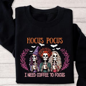 Op de afbeelding: Zwarte sweatshirt met een afbeelding van drie skeletheksen die koffiekopjes vasthouden, met de tekst "Hocus Pocus" en "I Need Coffee To Focus". Het ontwerp bevat vleermuizen en een bloemenkrans.