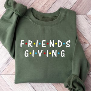 Pode incluir: Um moletom verde-oliva com o texto "FRIENDS GIVING" em branco, com pontos coloridos. O moletom está dobrado sobre uma superfície de madeira marrom clara e branca.