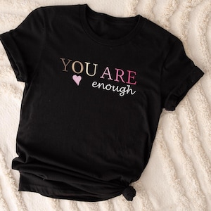Op de afbeelding: Zwart T-shirt met de tekst "YOU ARE enough" in roze, goud en witte letters met een roze hart.
