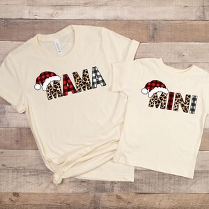 May include: Two cream-colored t-shirts with a Christmas theme. The larger shirt says "MAMA" in red, black, and white buffalo plaid, leopard print, and a red Santa hat. The smaller shirt says "MINI" in the same design.