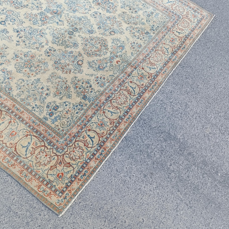 May include: Cream-colored area rug with an intricate floral pattern in shades of blue and red. The rug features a detailed border with a repeating design. The rug is placed on a gray surface.