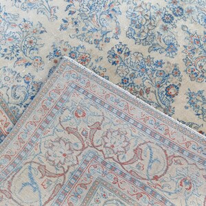 May include: An overhead shot of a vintage rug with a floral pattern. The rug features a cream-colored background with blue and red floral designs. The rug is folded over, revealing the fringed edge and detailed border.