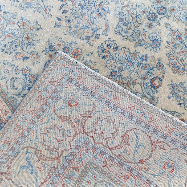 May include: An overhead shot of a vintage rug with a floral pattern. The rug features a cream-colored background with blue and red floral designs. The rug is folded over, revealing the fringed edge and detailed border.