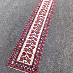 May include: A rectangular runner rug with a central design of red floral motifs on a cream background. The rug has a red border with geometric patterns and fringe on the ends. The rug is approximately 8 feet long.