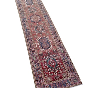 Puede incluir: Una alfombra pasillera larga y rectangular con un fondo rojo intenso y una serie de medallones geométricos repetidos en tonos azul, verde azulado y crema. El borde muestra patrones intrincados en azul y beige, con flecos en los extremos.