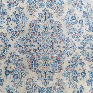 May include: Close-up of a vintage rug with a cream-colored background and an intricate floral pattern. The design features blue and coral-colored flowers and leaves, creating a symmetrical and detailed aesthetic. The rug's texture appears soft and worn.