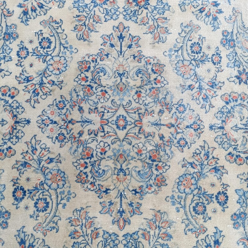 May include: Close-up of a vintage rug with a cream-colored background and an intricate floral pattern. The design features blue and coral-colored flowers and leaves, creating a symmetrical and detailed aesthetic. The rug's texture appears soft and worn.
