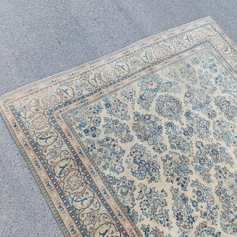 May include: An antique area rug with a cream-colored background and intricate floral patterns in shades of blue, brown, and beige. The rug features a detailed border design and a distressed, vintage appearance.