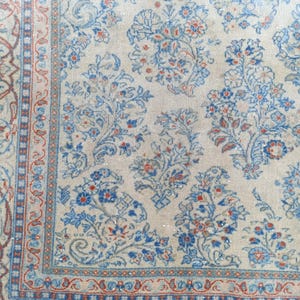 May include: Close-up of a beige rug with a floral pattern in blue and red. The rug has a detailed border with a repeating design. The floral pattern includes stylized flowers and leaves, creating a classic, ornate look. The rug appears to be made of a soft material.