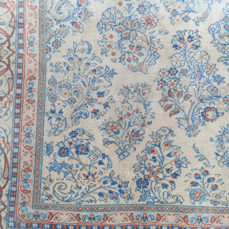 May include: Close-up of a beige rug with a floral pattern in blue and red. The rug has a detailed border with a repeating design. The floral pattern includes stylized flowers and leaves, creating a classic, ornate look. The rug appears to be made of a soft material.