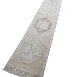 May include: A long, narrow runner rug featuring a neutral color palette of beige, cream, and soft gray. The design includes a series of repeating geometric diamond-shaped medallions centered along the length, each containing intricate linear patterns.