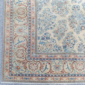 May include: Close-up of a beige rug with a detailed floral pattern in blue and red. The rug features a border with intricate designs and a central field with floral motifs, showcasing a vintage aesthetic.