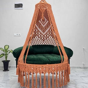 Puede incluir: Una silla colgante de macramé naranja con un cojín verde. La silla tiene un dosel en forma de cono y flecos decorativos. Está suspendida de una cadena de metal contra una pared blanca.