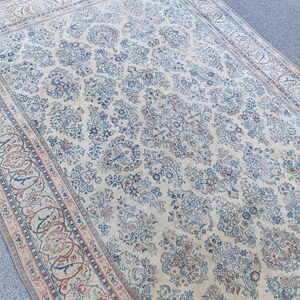May include: A large, rectangular rug with an intricate floral pattern in shades of cream, blue, and red. The rug features a central field of floral motifs and a detailed border. The overall design is traditional and ornate.