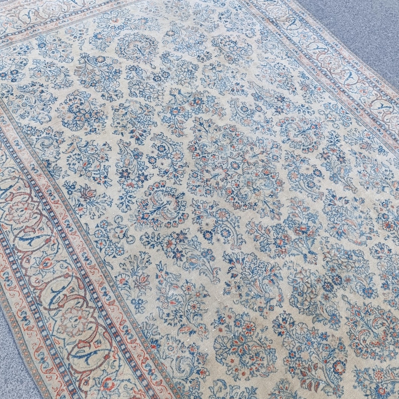 May include: A large, rectangular rug with an intricate floral pattern in shades of cream, blue, and red. The rug features a central field of floral motifs and a detailed border. The overall design is traditional and ornate.