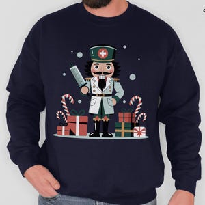 May include: Navy blue sweatshirt featuring a cartoon nutcracker dressed as a medical professional, holding a syringe. The design includes candy canes, wrapped gifts, and a cross symbol on the hat. The CozyCat Closet logo is in the upper right corner.