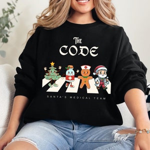 Op de afbeelding: Zwart sweatshirt met een witte afbeelding van een kerstboom, een fles medicijn, een peperkoekman en de kerstman die over een wit-zwarte gestreepte zebrapad lopen. De tekst "The Code" staat boven de afbeelding en "Santa's Medical Team" eronder.