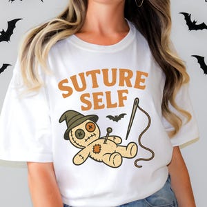 Könnte beinhalten: Weißes T-Shirt mit dem Text "SUTURE SELF" in Orange. Das Shirt zeigt eine Cartoon-Voodoo-Puppe mit Nadel und Faden und eine kleine Fledermaus. Schwarze Fledermaus-Silhouetten befinden sich im Hintergrund.