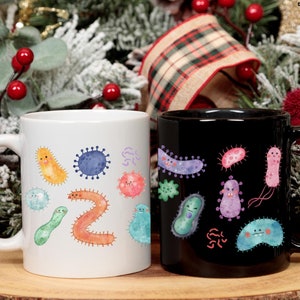 Könnte beinhalten: Zwei Keramikbecher, einer weiß und einer schwarz, jeweils mit farbenfrohen Illustrationen verschiedener Mikroorganismen verziert. Der weiße Becher zeigt eine Sammlung lächelnder Bakterien, während der schwarze Becher eine andere Reihe von Mikroben zeigt. Die Becher haben ein glänzendes Finish.