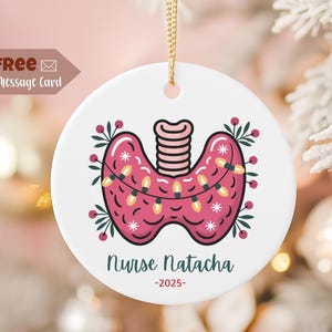 Może przedstawiać: Biała ceramiczna ozdoba świąteczna z różowym wzorem tarczycy, ozdobiona łańcuchem świetlnym i kwiatowymi akcentami. Ozdoba zawiera napis "Nurse Natacha" i rok "-2025". Do zawieszenia dołączony jest złoty sznurek.