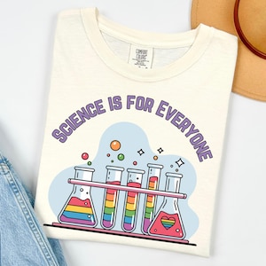 May include: Off-white t-shirt with the slogan "SCIENCE IS FOR EVERYONE" in purple. The design shows rainbow-coloured liquids in laboratory flasks and test tubes. The shirt is made of a soft material.
