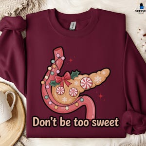 May include: Burgundy sweatshirt with a whimsical illustration of a pancreas decorated with Christmas lights, candy, and a bow. The text "Don't be too sweet" is printed below the graphic. CozyCat Closet logo is in the top right corner.