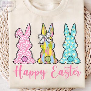 May include: A beige sweatshirt with three bunny silhouettes. The bunnies are decorated with different patterns, including leopard print, floral, and geometric designs. The bunnies are arranged in a row, and the text "Happy Easter" is printed below them in pink.