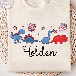 May include: Cream-colored sweatshirt featuring a row of cartoon dinosaurs in blue and red, with fireworks and stars. The name "Holden" is written in black script below the dinosaurs. The upper left corner has the text "ORIGINAL DESIGNER".