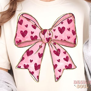 May include: A pink bow with gold glitter accents and a pattern of pink hearts. The bow is on a cream-colored t-shirt.
