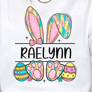 May include: White sweatshirt with a pink and yellow bunny ear design, the name "RAELynn" in black text, and colorful Easter eggs.
