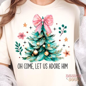 May include: A white t-shirt with a watercolor illustration of a Christmas tree with a pink bow, gold ornaments, and green leaves. The text "Oh Come, Let Us Adore Him" is printed below the tree.