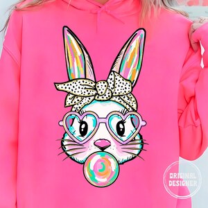 May include: A pink hoodie featuring a graphic of a white bunny wearing heart-shaped sunglasses and a polka dot headband. The bunny is blowing a bubblegum bubble.